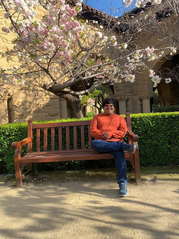 Tejas sitting on a wooden bench under a blossoming cherry tree at Stanford